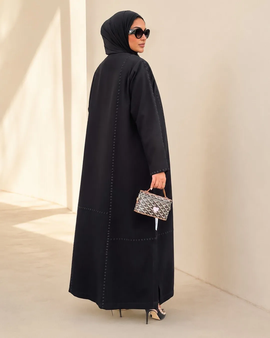 Back view of a black abaya with raised stitch lines against a sand wall