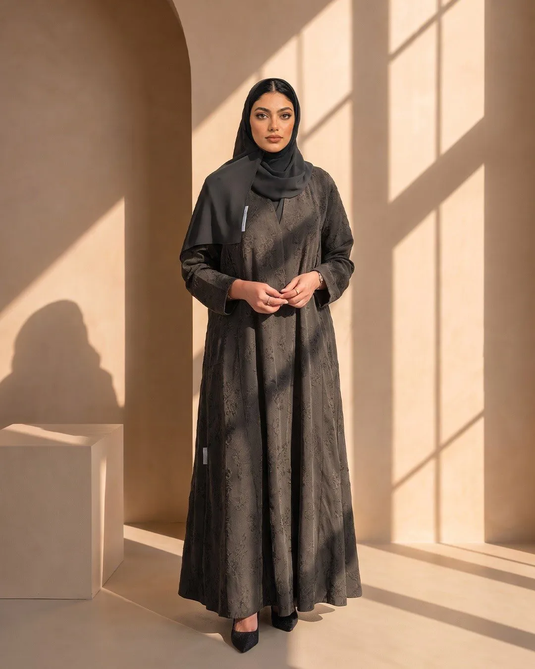 Model in a charcoal floral jacquard abaya standing in a sunlit room with an arched doorway