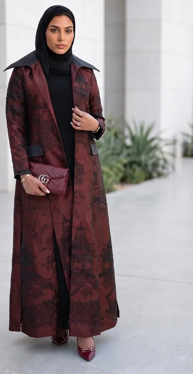 Model in a burgundy brocade coat abaya with a wide black collar in a stone colonnade
