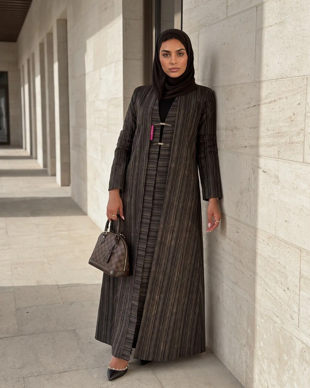 Model in a brown striped layered abaya leaning on a travertine wall